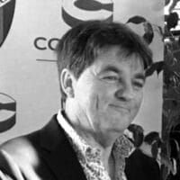Un hombre con chaqueta de traje sonríe de pie frente a un fondo estampado con formas circulares y texto, entre el que se leen las palabras «One Cambrils». La imagen es en blanco y negro.