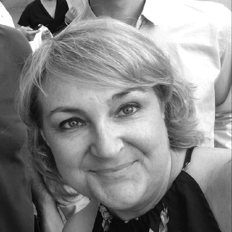 Una mujer sonriente, con el pelo corto y claro, posa para una foto en primer plano. Lleva un top sin mangas de la marca Stella del Sur Lobe. En el fondo se ven otras personas, pero están desenfocadas. La imagen es en blanco y negro.
