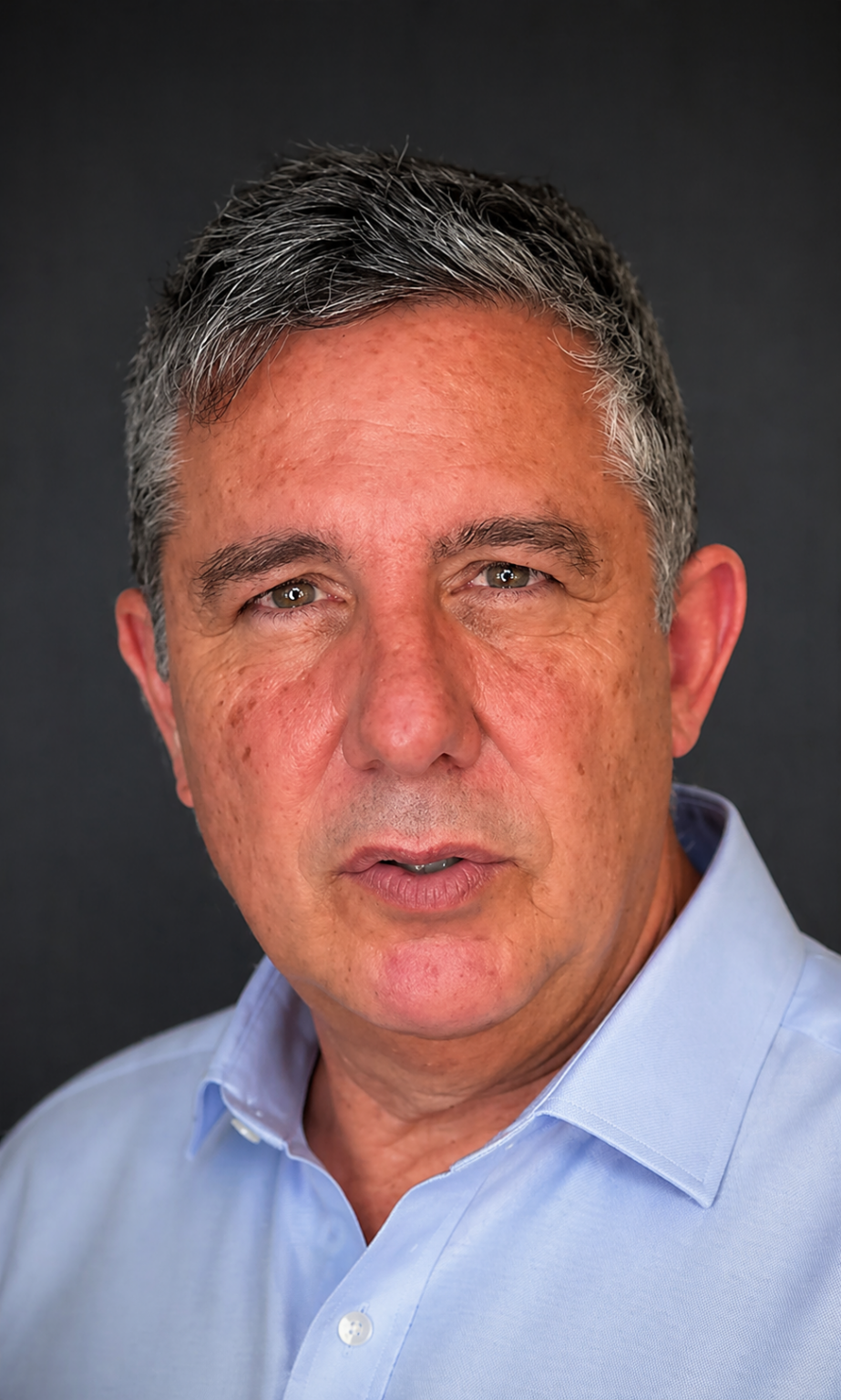 Hombre de mediana edad, con el pelo corto y canoso, que lleva una camisa de cuello azul claro y mira directamente a la cámara con expresión neutra sobre un fondo oscuro, lo que recuerda a un retrato del dossier Gayarre.